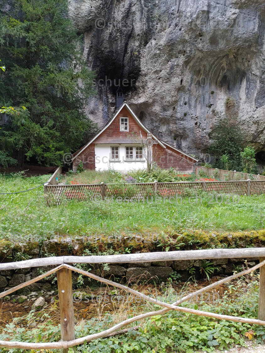 Hermitage of St. Verena in front of a high rock face. There is a small garden in front of the house.